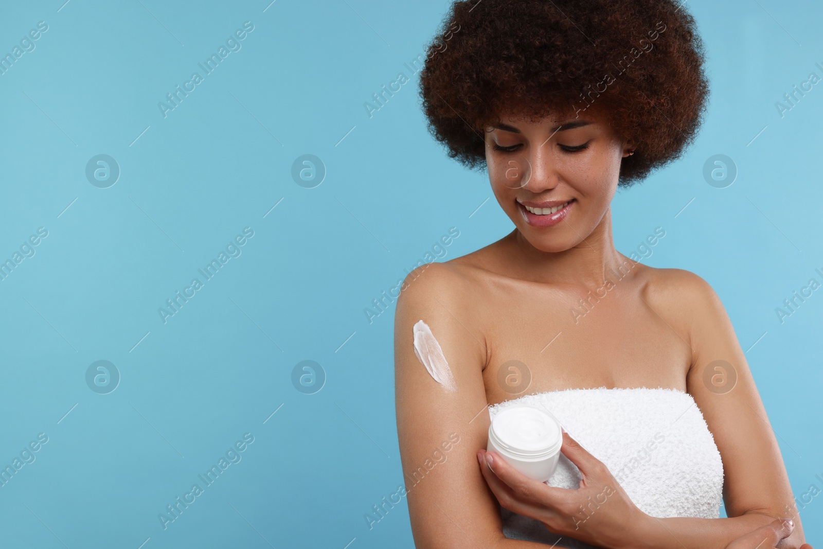 Photo of Beautiful young woman applying body cream onto arm on light blue background, space for text