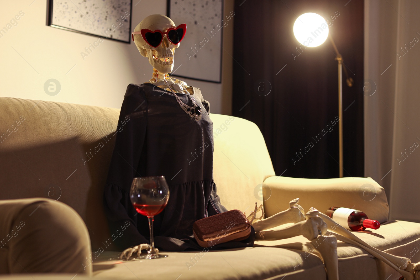 Photo of Skeleton in dress with wine sitting on sofa at home