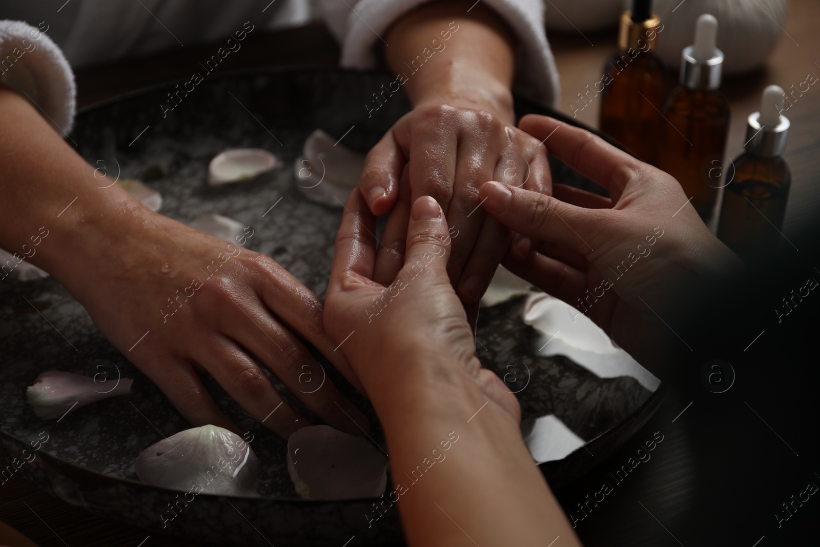 Photo of Woman receiving hand massage in spa salon, closeup. Bowl of water and flower petals on table