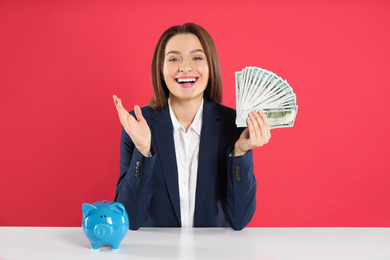 Photo of Young woman with money and piggy bank at table on crimson background