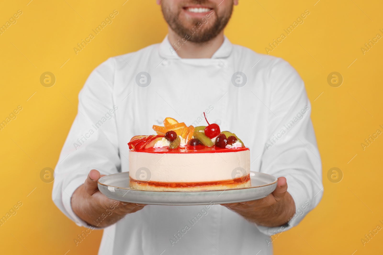Photo of Happy professional confectioner in uniform holding delicious cake on yellow background, closeup