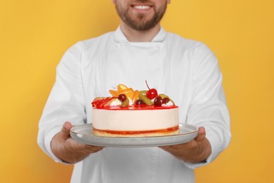 Photo of Happy professional confectioner in uniform holding delicious cake on yellow background, closeup