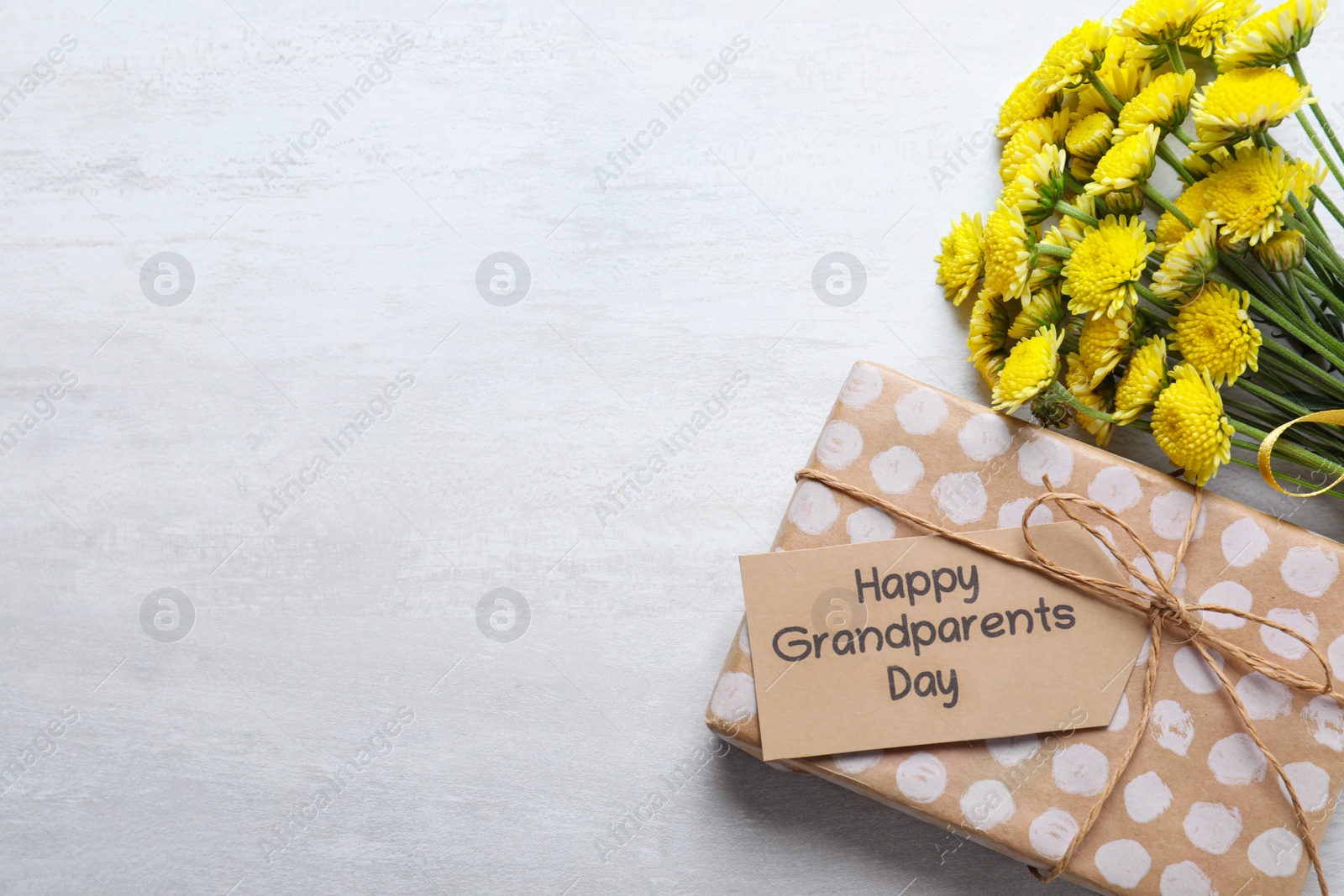 Photo of Beautiful yellow flowers, gift box and tag with phrase Happy Grandparents Day on light background, flat lay. Space for text