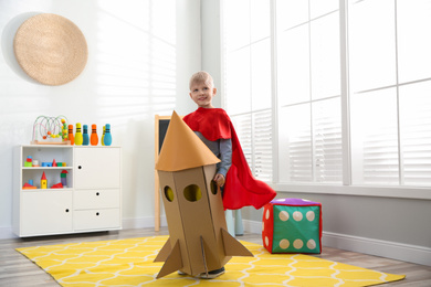 Little child in red cape playing with rocket made of cardboard box at home