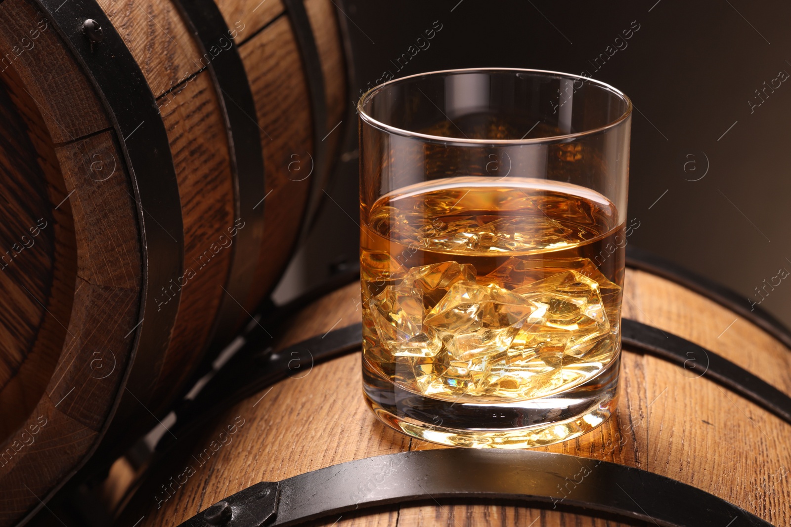 Photo of Whiskey with ice cubes in glass on wooden barrel against dark background, closeup