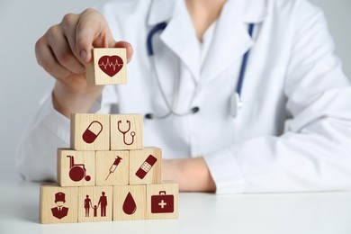 Image of Doctor building pyramid of wooden cubes with different icons at white table, closeup. Insurance concept