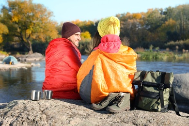 Photo of Couple of campers in sleeping bags sitting on rock near pond