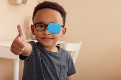 Photo of African American boy with eye patch on glasses showing thumb up in room, space for text. Strabismus treatment