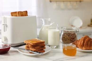 Photo of Making toasts for breakfast. Appliance, crunchy bread, honey, jam, milk and croissant on white marble table in kitchen