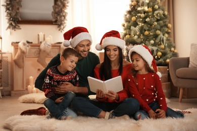 Photo of Happy family in Santa hats reading book on floor at home