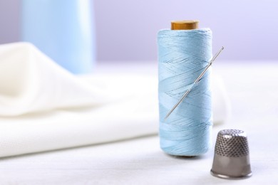 Spool of light blue sewing thread with needle and thimble on white table, space for text