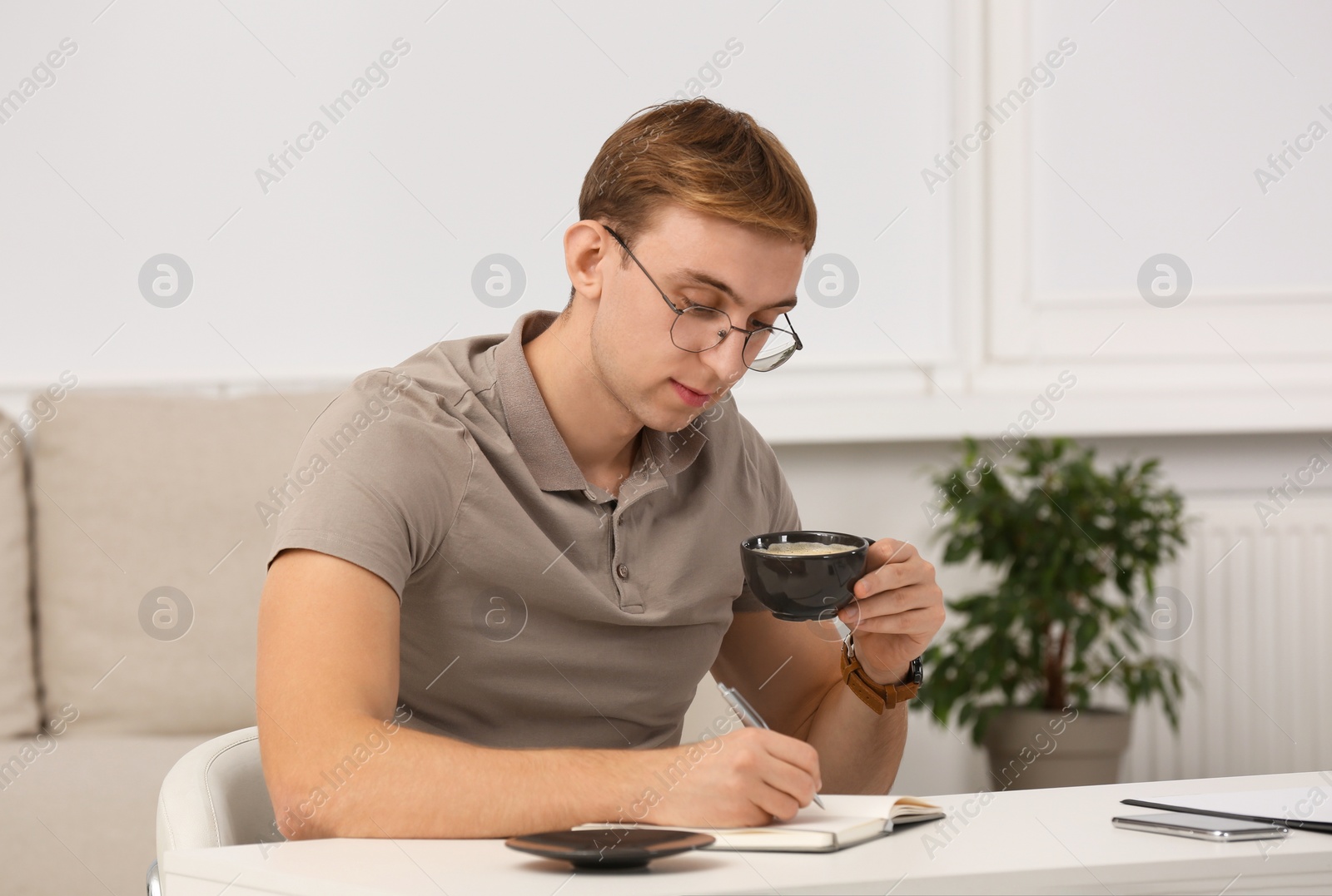 Photo of Young man with cup of coffee writing in notebook at table indoors