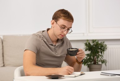 Photo of Young man with cup of coffee writing in notebook at table indoors