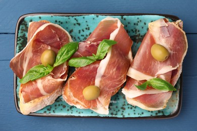 Photo of Tasty sandwiches with cured ham and basil leaves on blue wooden table, top view