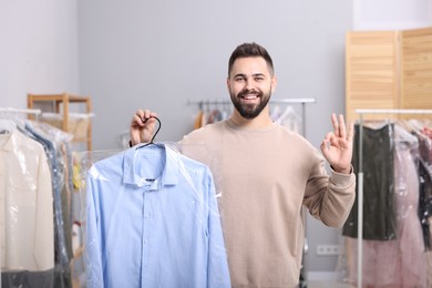 Dry-cleaning service. Happy man holding hanger with shirt in plastic bag and showing ok gesture indoors