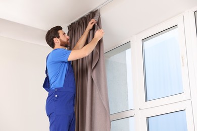 Worker in uniform hanging window curtain indoors