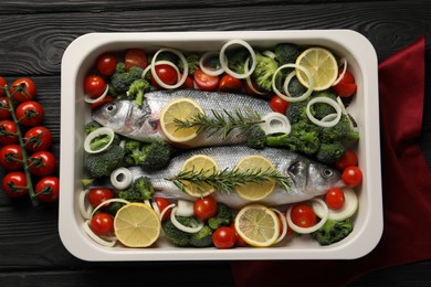 Raw fish with lemon in baking dish and vegetables on black wooden table, flat lay