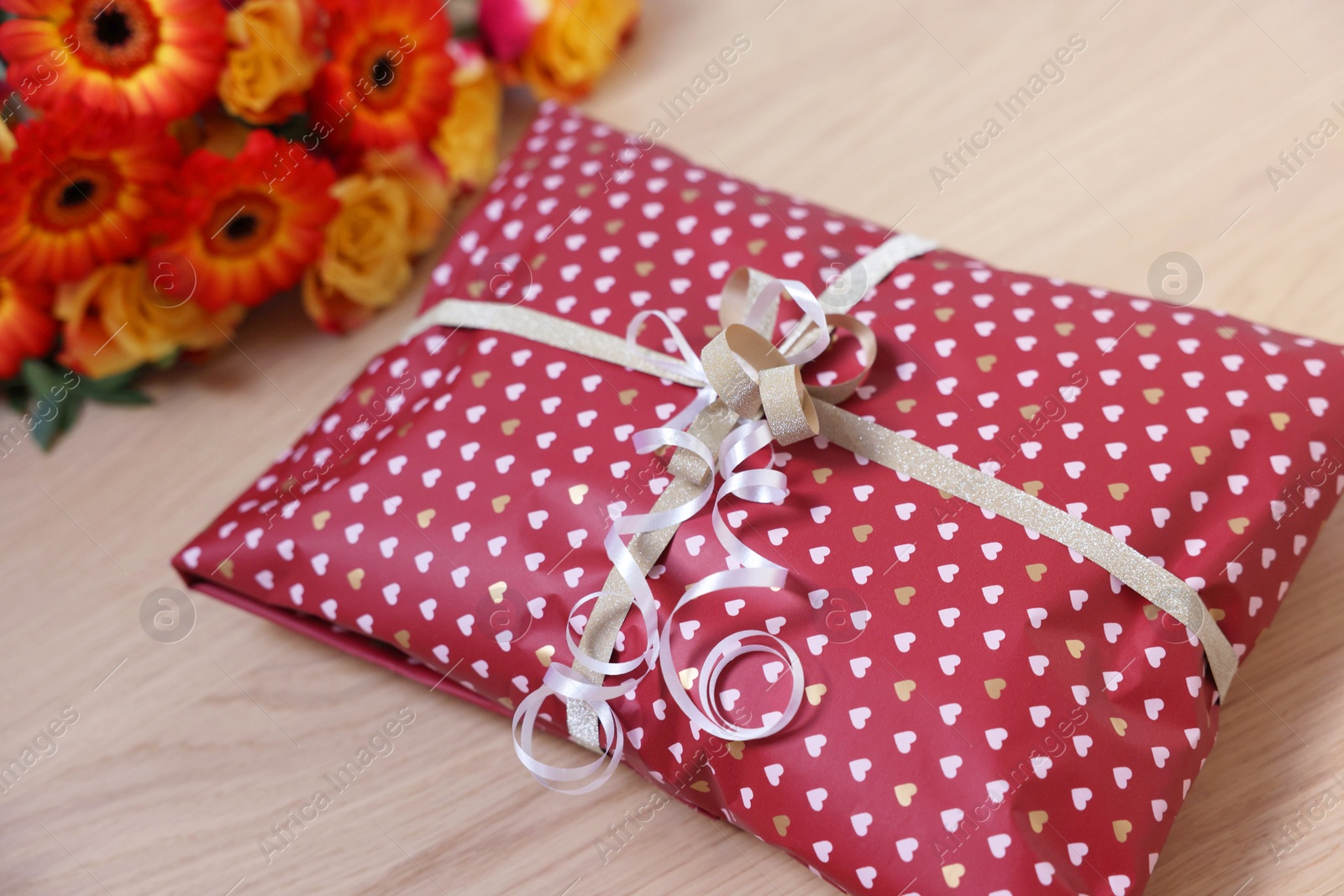 Photo of Parcel wrapped in heart patterned paper and beautiful flowers on wooden table, closeup