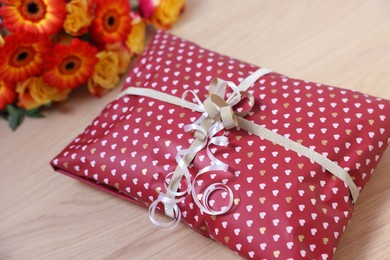 Parcel wrapped in heart patterned paper and beautiful flowers on wooden table, closeup