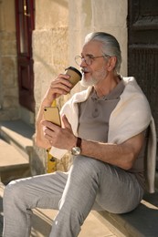 Photo of Handsome senior man sitting on doorstep with smartphone and drinking coffee outdoors