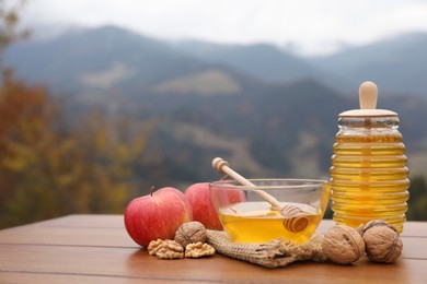 Photo of Fresh aromatic honey, apples and walnuts on wooden table against mountain landscape. Space for text