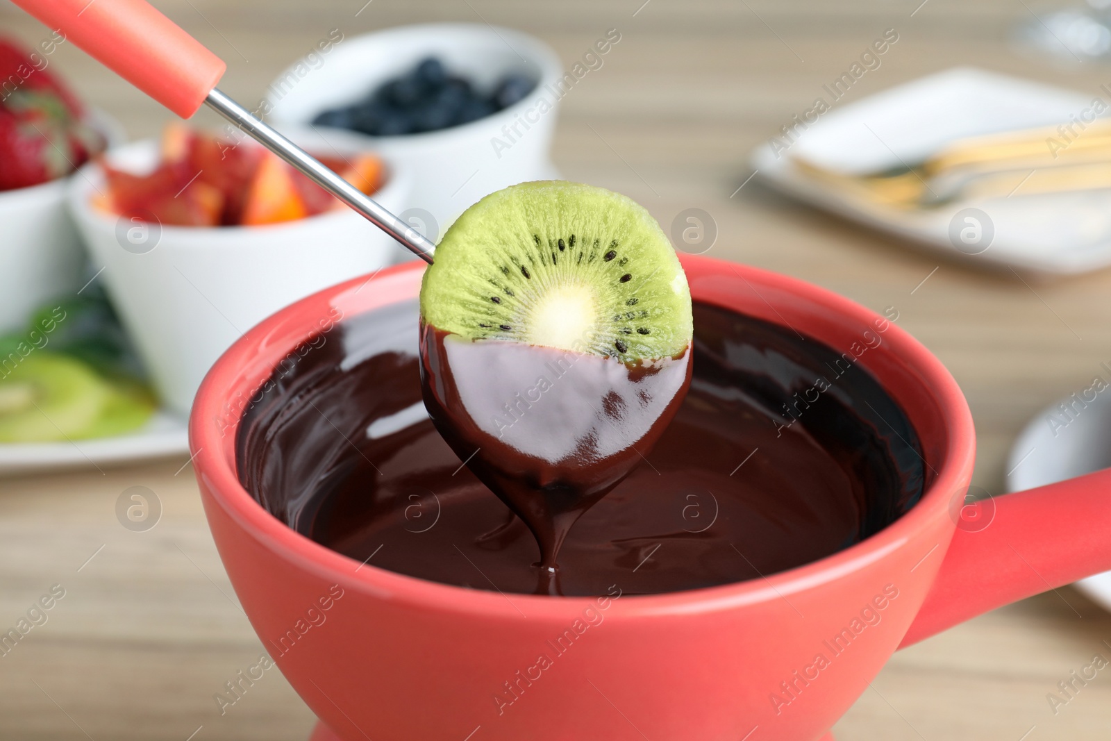 Photo of Dipping kiwi slice into pot with chocolate fondue on table, closeup