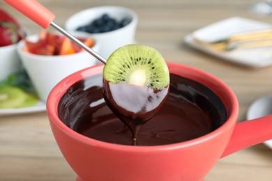 Dipping kiwi slice into pot with chocolate fondue on table, closeup