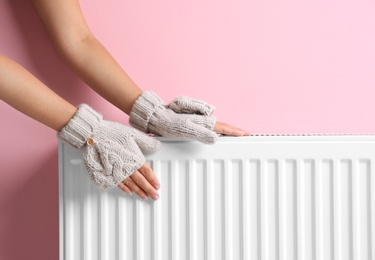 Woman in mittens warming hands on heating radiator near color wall
