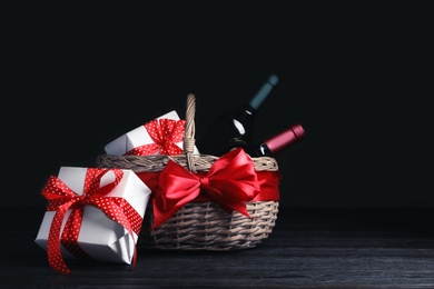 Photo of Festive basket with bottles of wine and gifts on table against dark background. Space for text