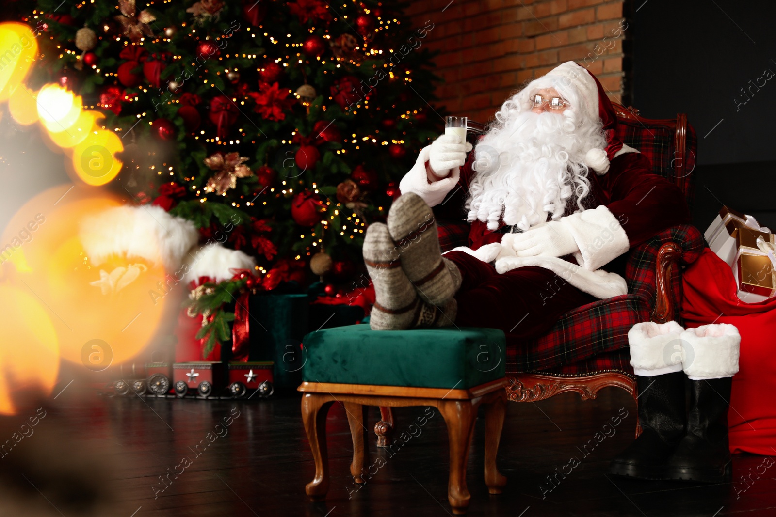 Photo of Santa Claus with glass of milk resting in armchair near Christmas tree