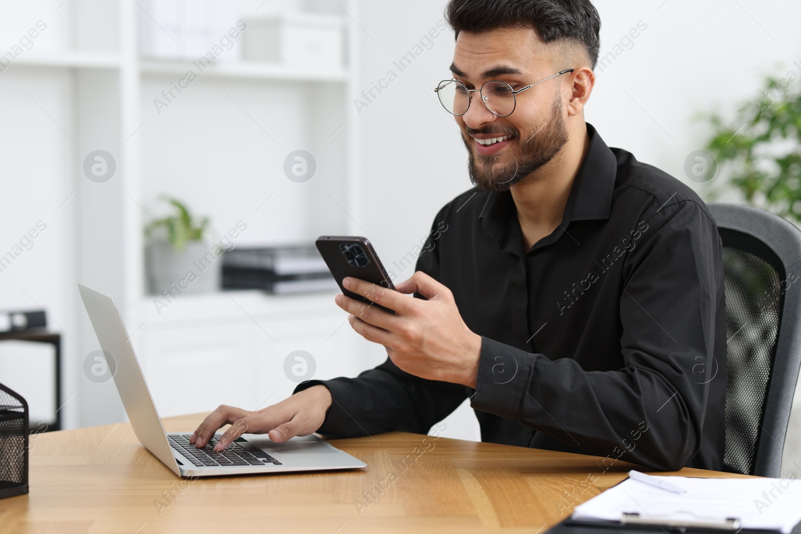 Photo of Handsome young man using smartphone while working with laptop at wooden table in office