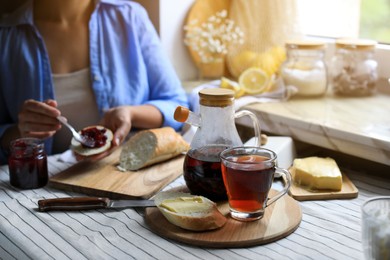Photo of Woman spreading jam onto bread at table indoors, focus on aromatic tea