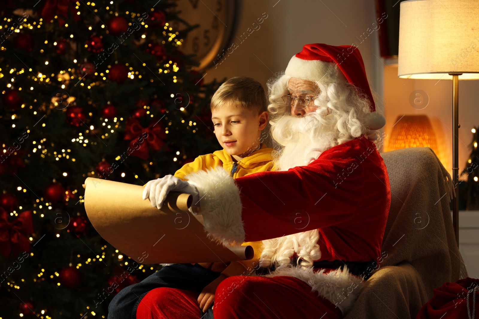 Photo of Santa Claus with little boy reading letter near Christmas tree in room