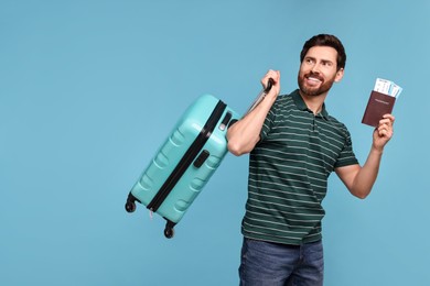 Smiling man with passport, suitcase and tickets on light blue background. Space for text