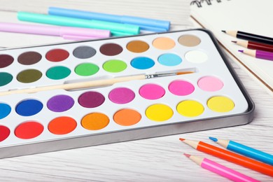 Watercolor palette with brush, colorful pencils and markers on white wooden table, closeup