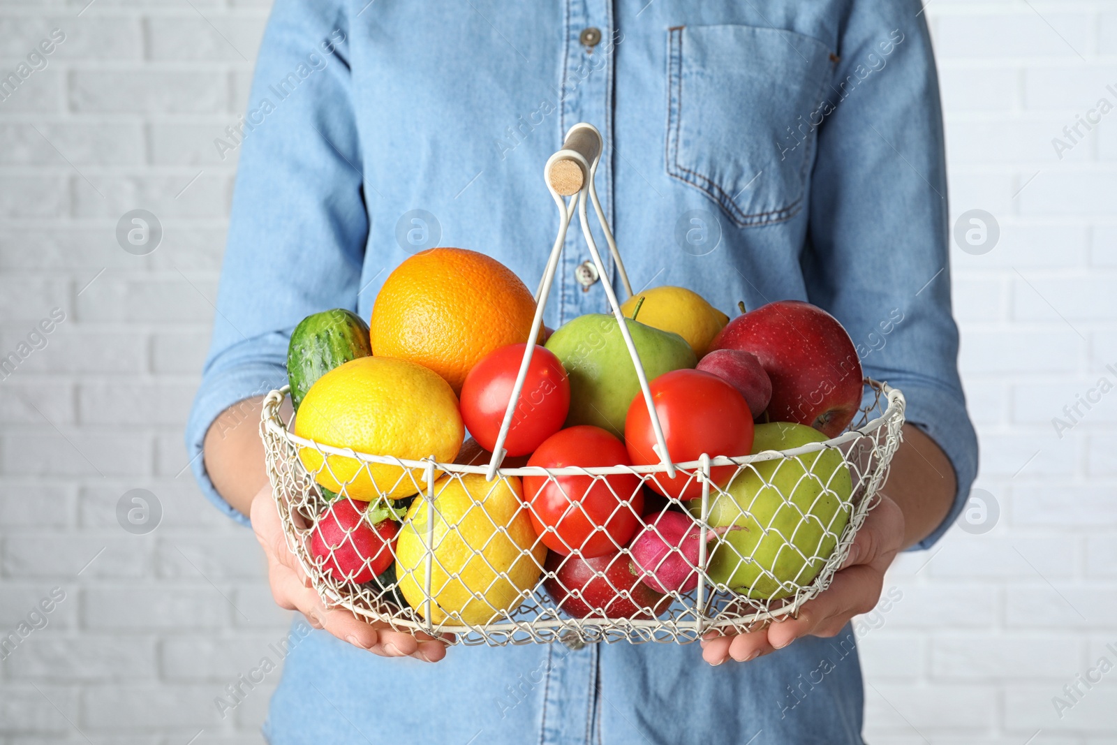 Photo of Woman holding basket with ripe fruits and vegetables near white brick wall, closeup