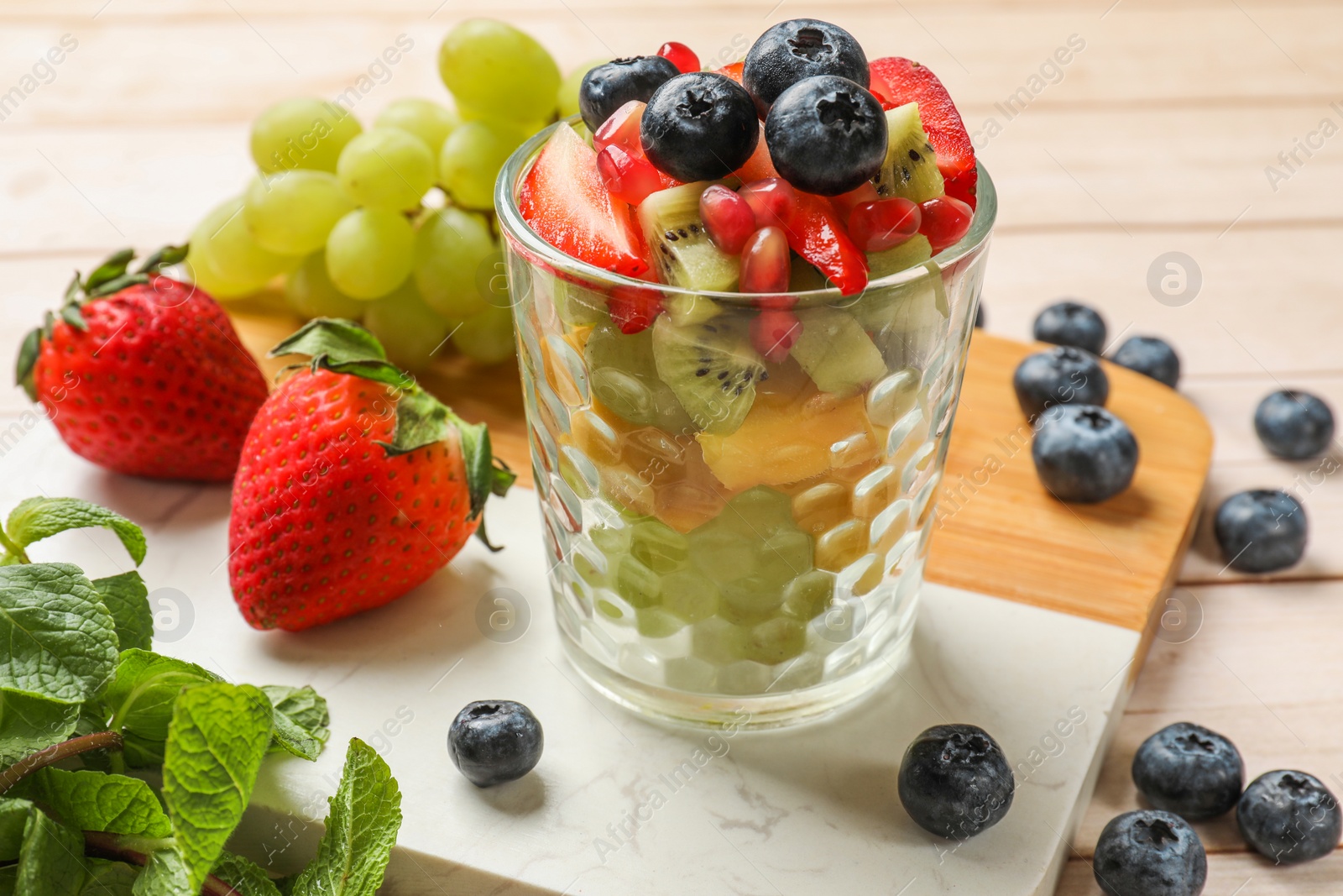 Photo of Healthy breakfast. Delicious fruit salad in glass and ingredients on light wooden table, closeup