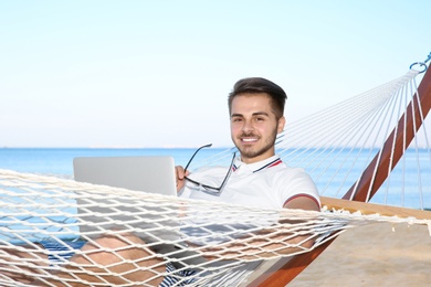 Young man with laptop resting in hammock at seaside