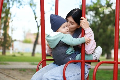 Mother holding her child in sling (baby carrier) on swing outdoors. Space for text