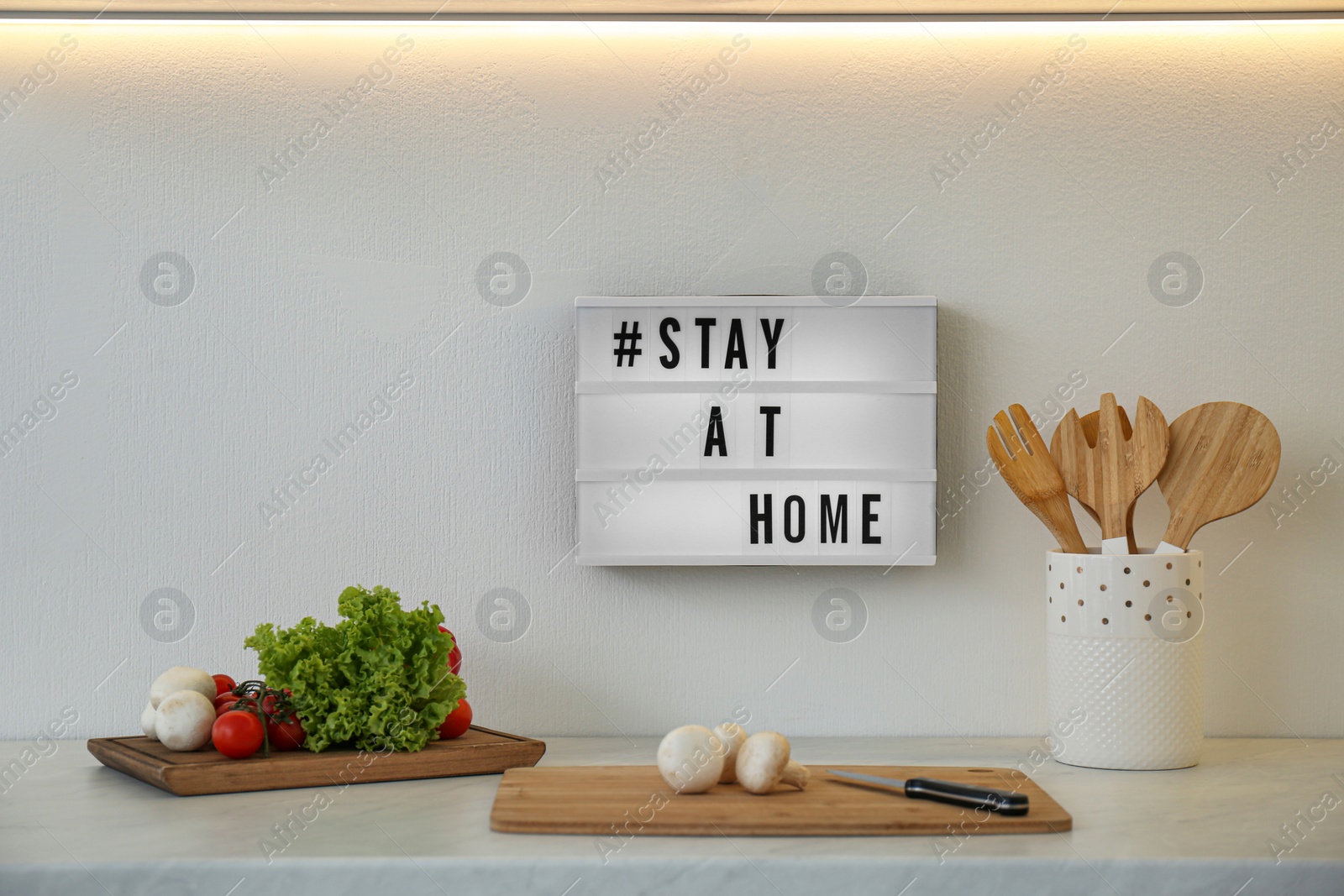 Photo of Fresh products, cooking utensils and lightbox with hashtag STAY AT HOME in kitchen. Message to promote self-isolation during COVID‑19 pandemic