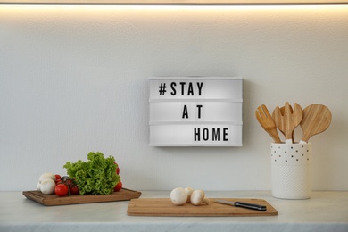 Fresh products, cooking utensils and lightbox with hashtag STAY AT HOME in kitchen. Message to promote self-isolation during COVID‑19 pandemic