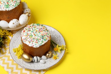 Traditional Easter cakes with sprinkles, painted eggs and beautiful spring flowers on yellow background, space for text