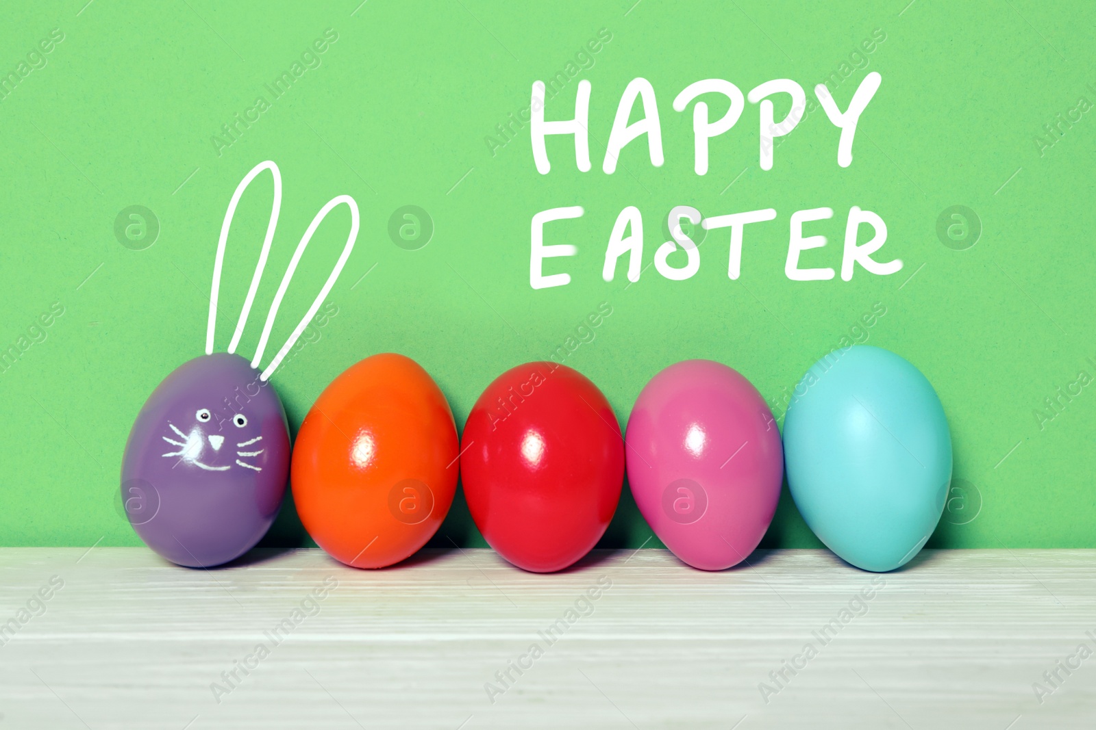 Image of Violet egg with drawn face and ears as Easter bunny among others on white wooden table against green background