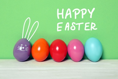 Image of Violet egg with drawn face and ears as Easter bunny among others on white wooden table against green background