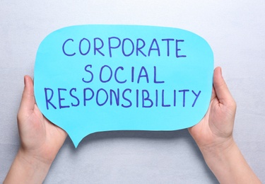 Photo of Woman holding speech balloon with phrase Corporate Social Responsibility on light grey background, top view