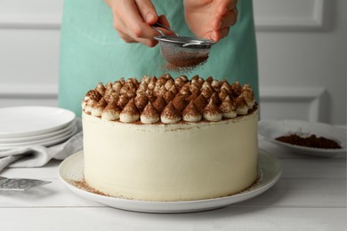 Photo of Woman dusting delicious tiramisu cake with cocoa powder at white wooden table, closeup