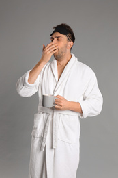 Young man in bathrobe yawning on grey background