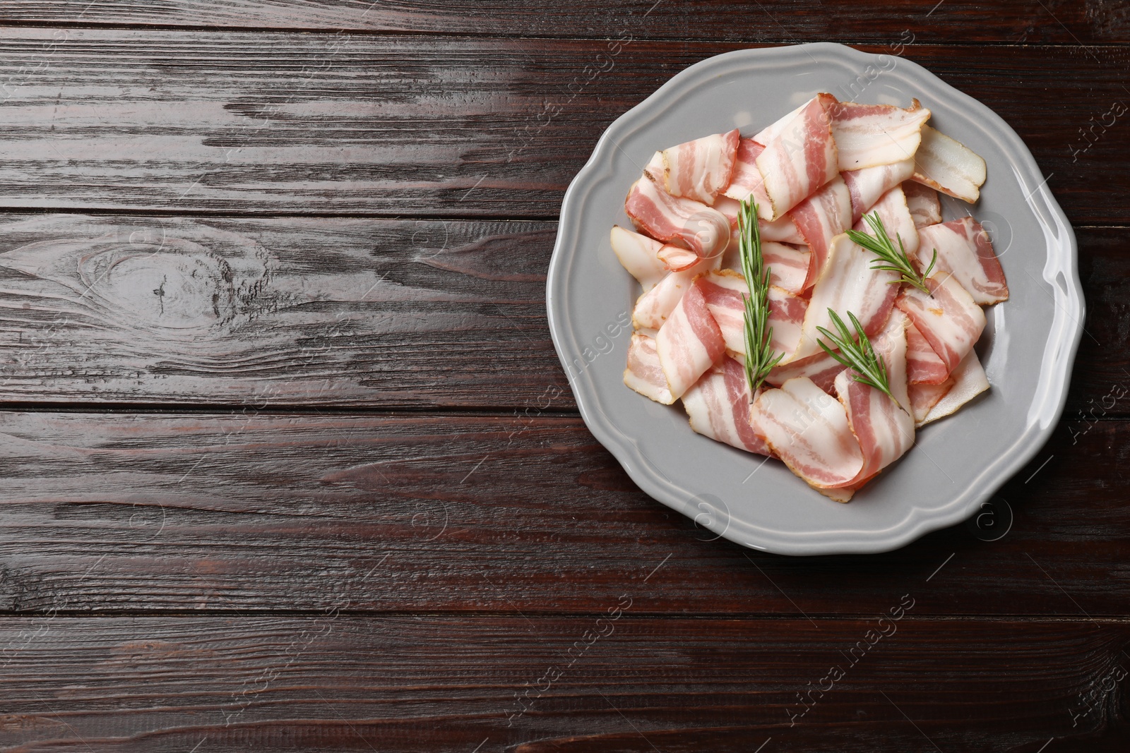 Photo of Slices of raw bacon and rosemary on dark wooden table, top view. Space for text