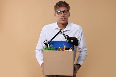 Unemployed young man with box of personal office belongings on beige background
