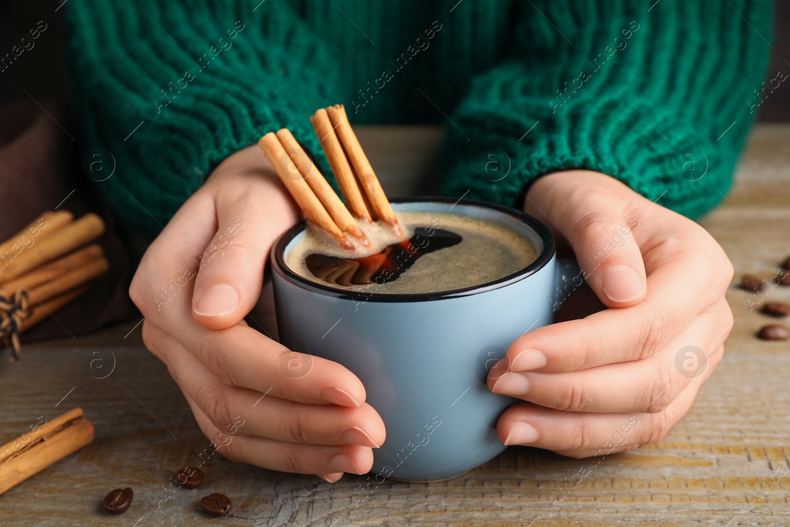 Photo of Woman holding cup of hot coffee with aromatic cinnamon at wooden table, closeup